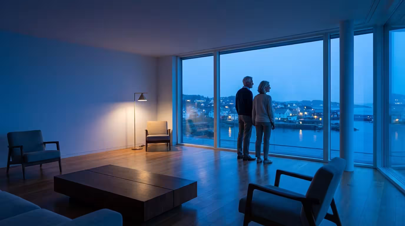 An older couple in a modern home look out at a coastal town at dusk.