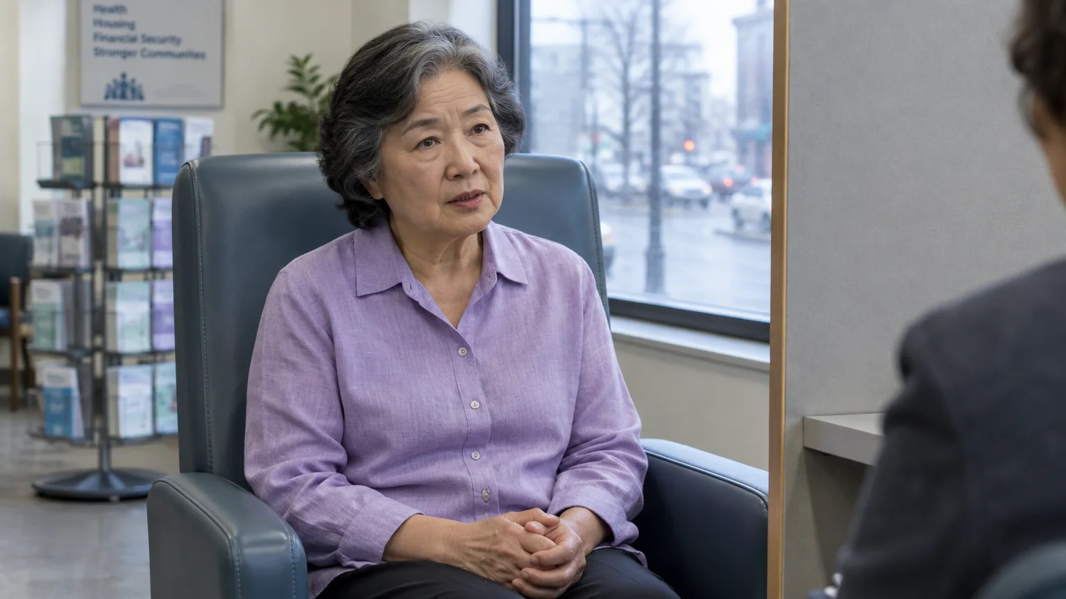 A mature Asian woman in a purple blouse sits in an armchair across from an interviewer in an office near a window.