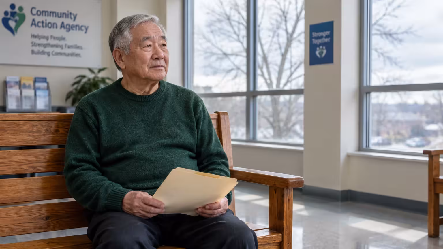 Senior Asian man in a green sweater holding a manila folder while seated on a wooden bench at a Community Action Agency.