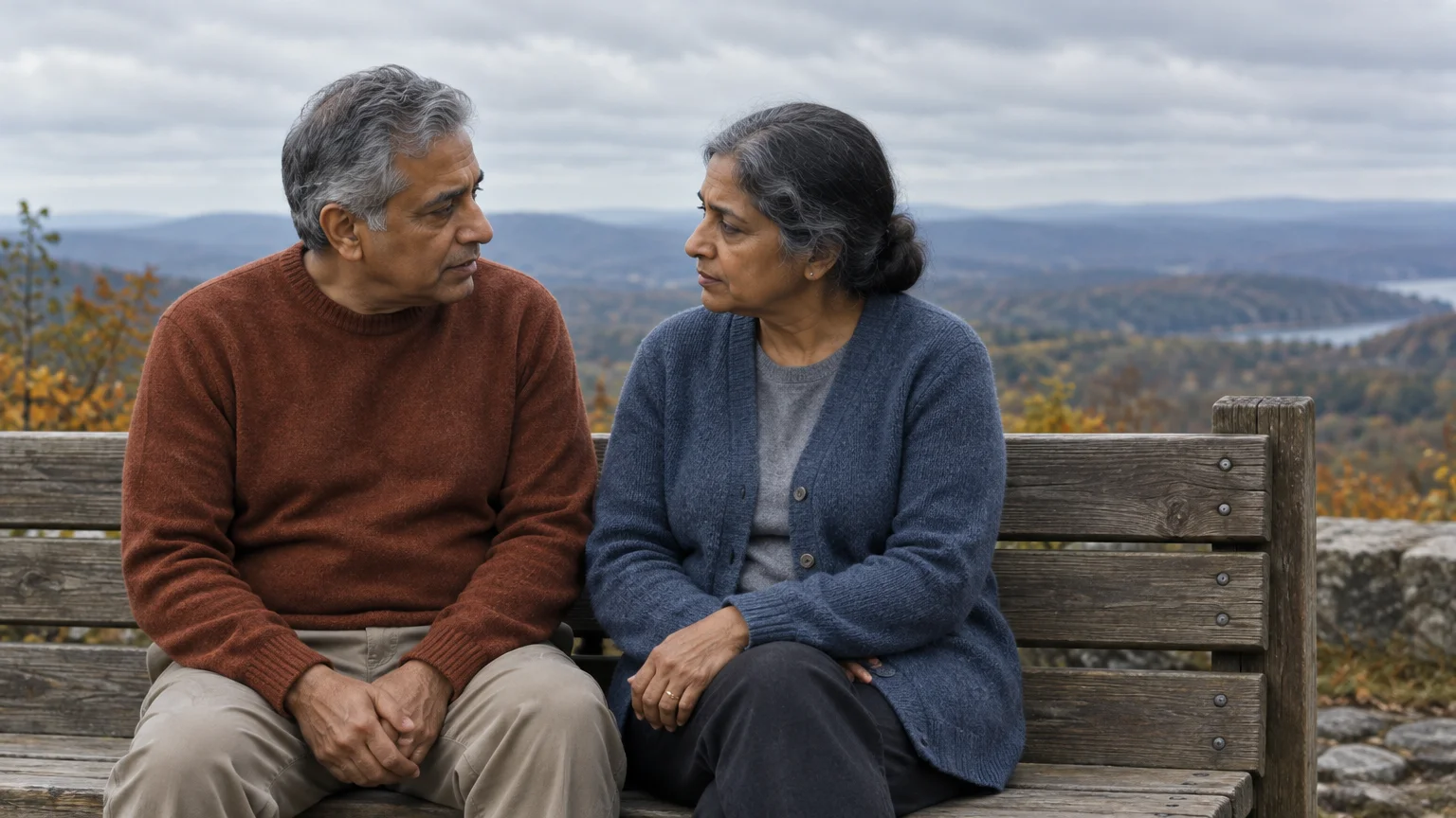 Mature South Asian man and woman sitting on a wooden bench outdoors, talking against a scenic mountain overlook.