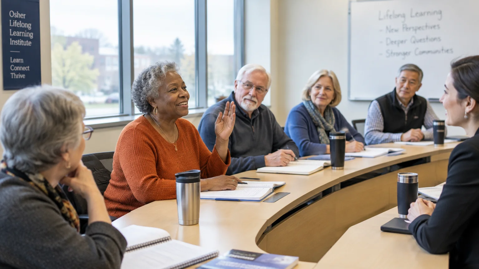 Older adults sit at a curved seminar table listening to an instructor, with one woman raising her hand to speak.