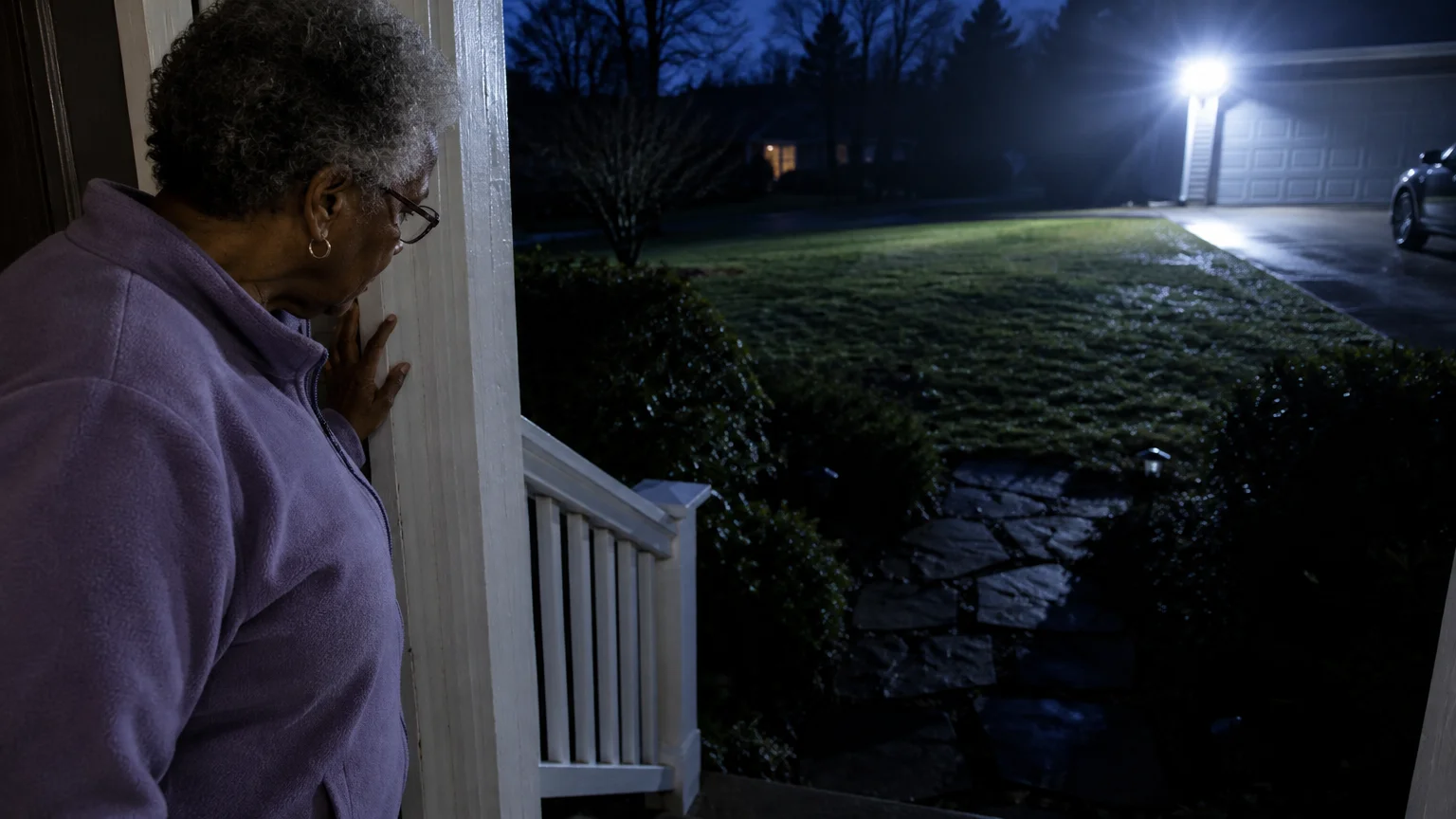 An older woman in glasses looks out from a porch at a dark stone walkway illuminated by a bright floodlight.