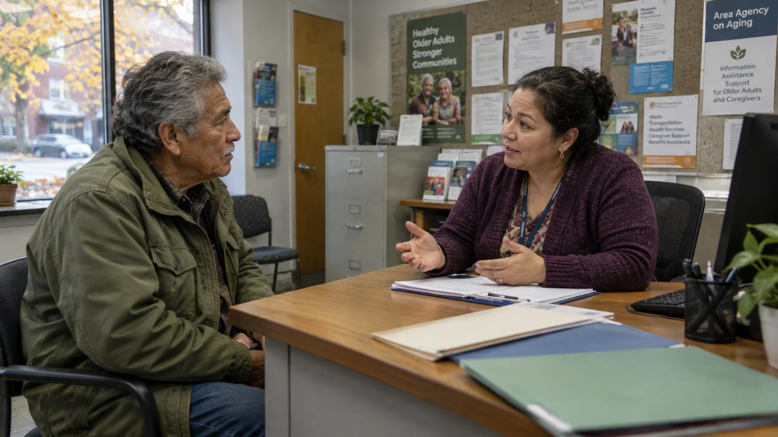 A woman sits at an office desk speaking with an older man seated across from her, with paperwork laid between them.