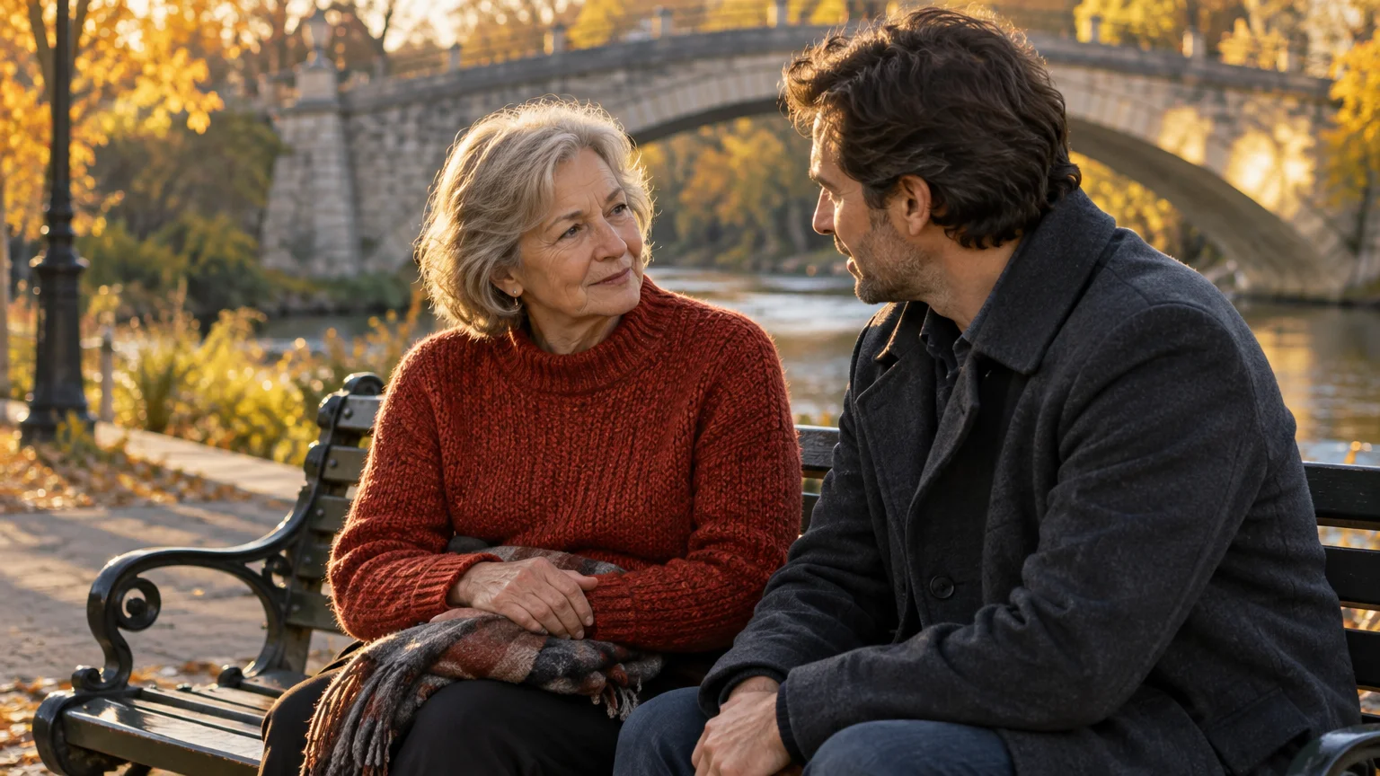 Older woman and man talking on a park bench with a stone arch bridge and river in the background during autumn.