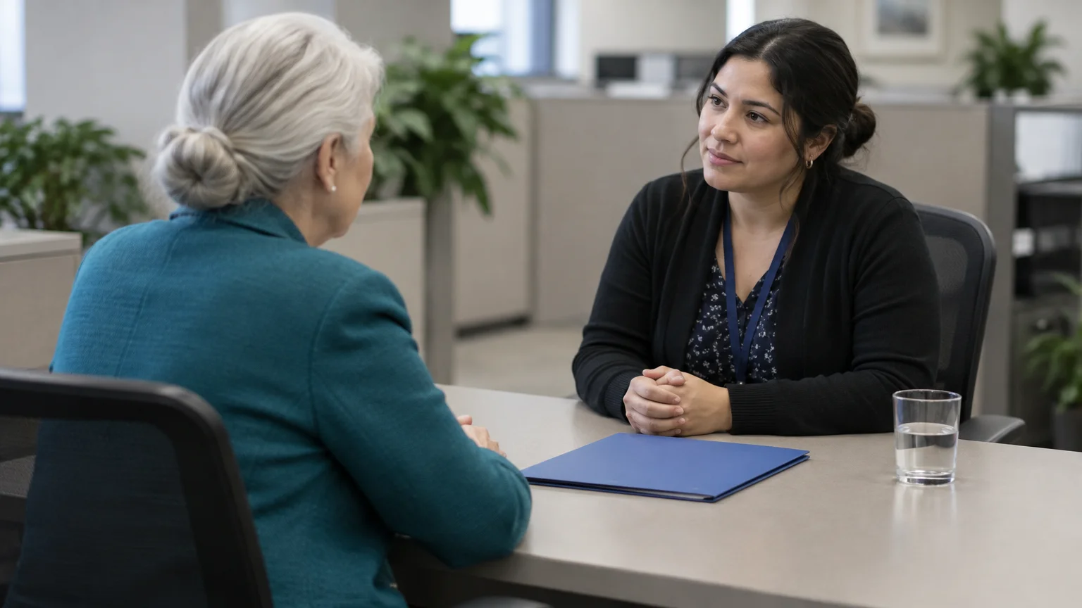 A woman with dark hair sits at an office desk opposite an older woman, with a blue folder between them.